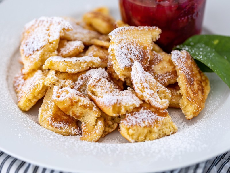 Bananen-Schmarrn mit Puderzucker und Zwetschgenkompott auf einem Teller.