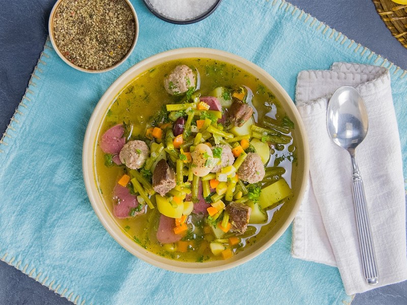 Ein Bohneneintopf mit gepökelter Schweinezunge auf einem blauen Tuch. Rechts daneben liegt ein Löffel auf einer weißen Serviette. Drumherum liegen einige Zutaten verteilt.