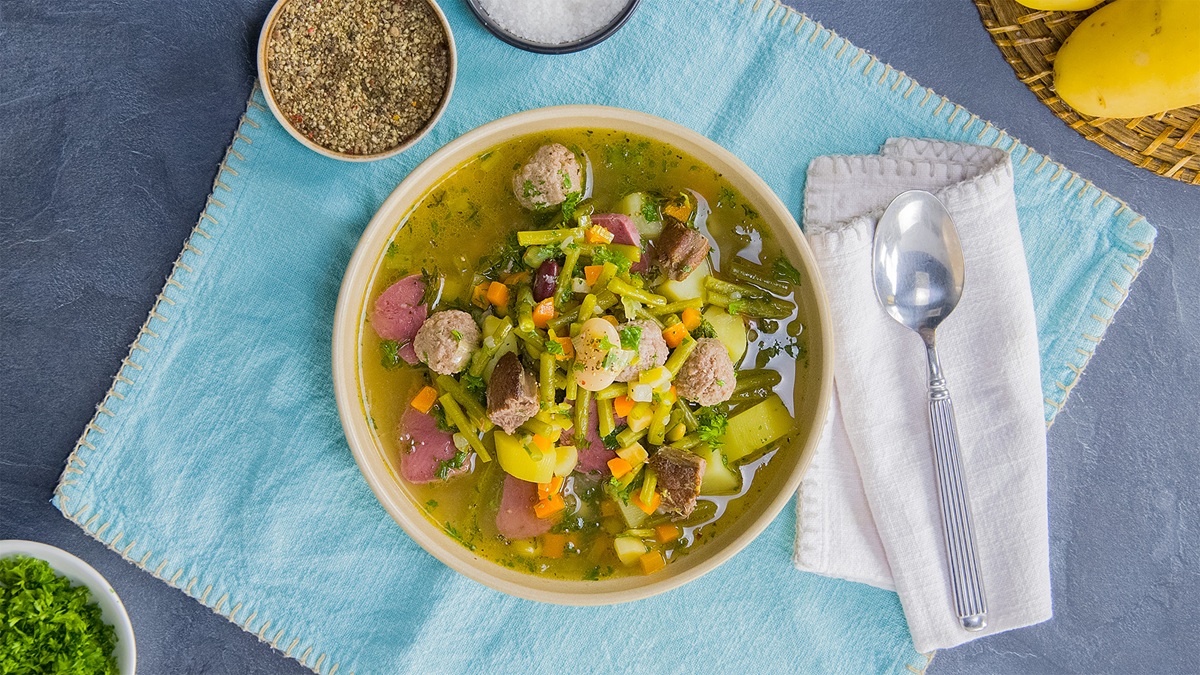 Ein Bohneneintopf mit gepökelter Schweinezunge auf einem blauen Tuch. Rechts daneben liegt ein Löffel auf einer weißen Serviette. Drumherum liegen einige Zutaten verteilt.