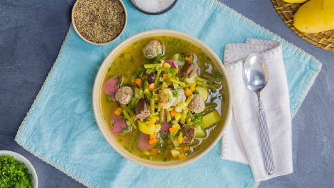 Ein Bohneneintopf mit gepökelter Schweinezunge auf einem blauen Tuch. Rechts daneben liegt ein Löffel auf einer weißen Serviette. Drumherum liegen einige Zutaten verteilt.