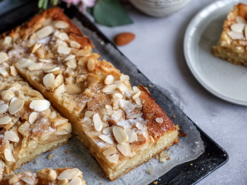 Ein angeschnittener Bratapfel-Butterkuchen auf einem Blech. Ein Stück auf einem Teller daneben.