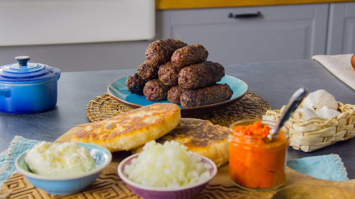 Ein Stapel Cevapcici auf einem Teller im Hintergrund. Vorne sind Fladenbrot und weitere Zutaten zu sehen.