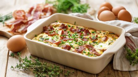 Eine Auflaufform mit Ei-Bacon-Schnitten, daneben rohe Eier und Kräuter.