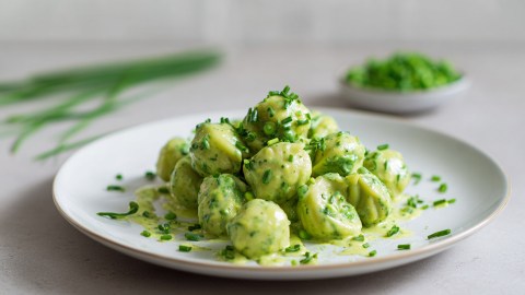 Ein Teller mit einer Portion Erbsen-Pasta mit Frischkäse-Senfsoße. Im Hintergrund kann man einen Bund Schnittlauch und eine Schale mit Erbsen erkennen.