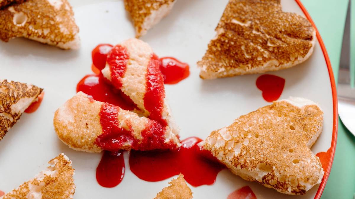 Ein Pancake in Herzform, der in der Mitte zerteilt ist und mit roter Fruchtsoße beträufelt ist. Das Ganze soll ein gebrochenes Herz symbolisieren. Drum herum liegen noch einige weitere kleine Pancakes in Herform.