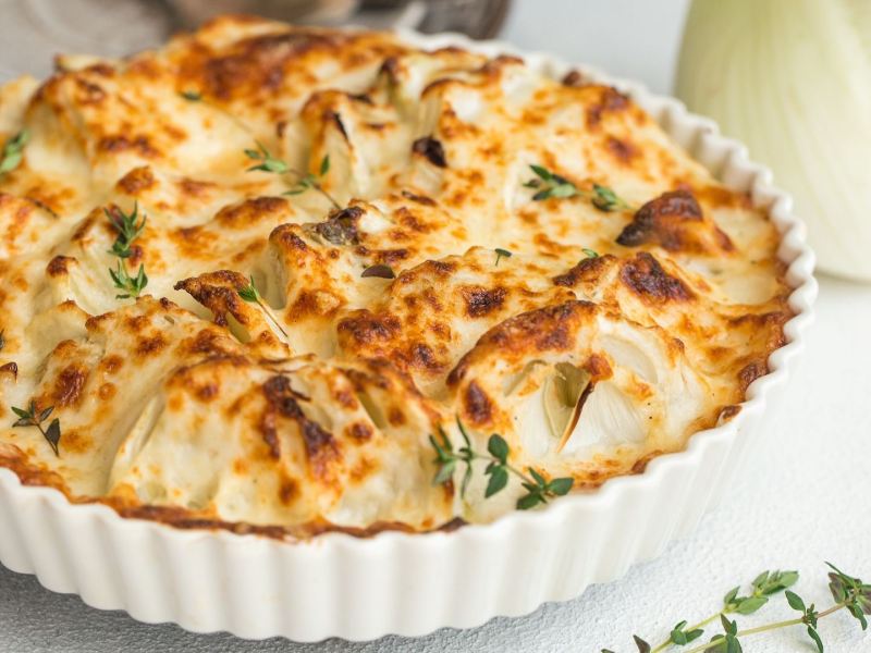 Eine Auflaufform mit Fenchelgratin, daneben eine Fenchelknolle.