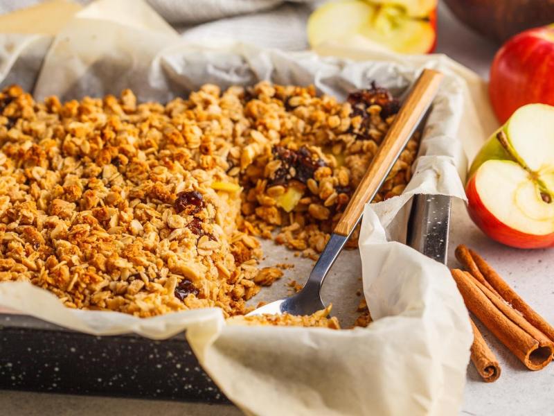 Gebackene Apfelkuchen-Haferflocken in einer Auflaufform, daneben eine Zimtstange und Äpfel.