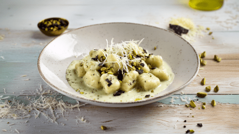 Gnocchi mit Pistaziencreme auf einem grauen Teller. Vorne links vor dem Teller liegt frisch geriebener Parmesan. Hinten links ein Schälchen mit Gewürzen. Rechts vom Teller liegen gehackte Pistazien.