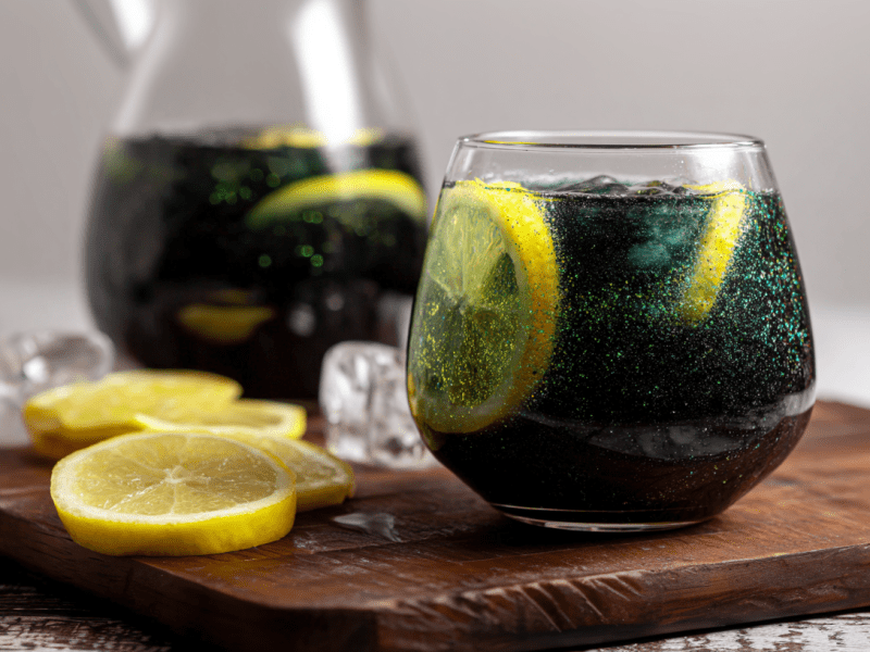 Ein Glas Hexen-Limonade steht auf einem Holzbrett. Daneben liegen einige Zitronenscheiben. Im Hintergrund ist noch eine Karaffe mit der Limo.