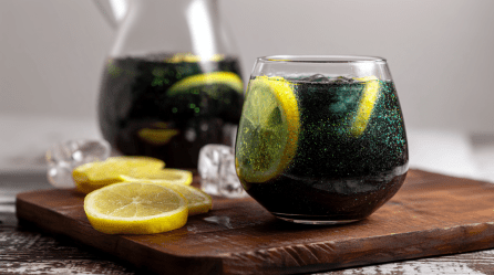 Ein Glas Hexen-Limonade steht auf einem Holzbrett. Daneben liegen einige Zitronenscheiben. Im Hintergrund ist noch eine Karaffe mit der Limo.