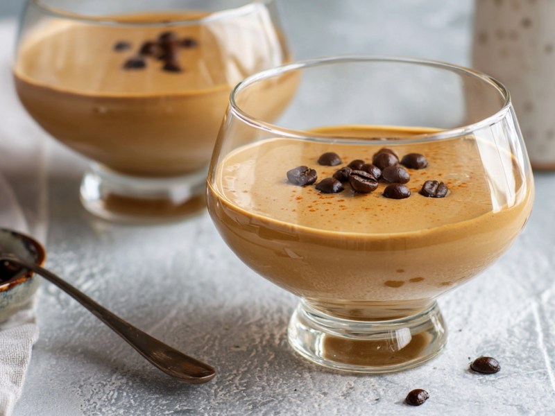 Zwei Dessertgläser mit Hopjesvla, einem holländischen Kaffee-Karamell-Pudding. Beide Puddings sind mit Kaffeebohnen garniert. Links daneben ist eine kleine Schale mit Kaffeebohnen und einem Löffel darin zu sehen.