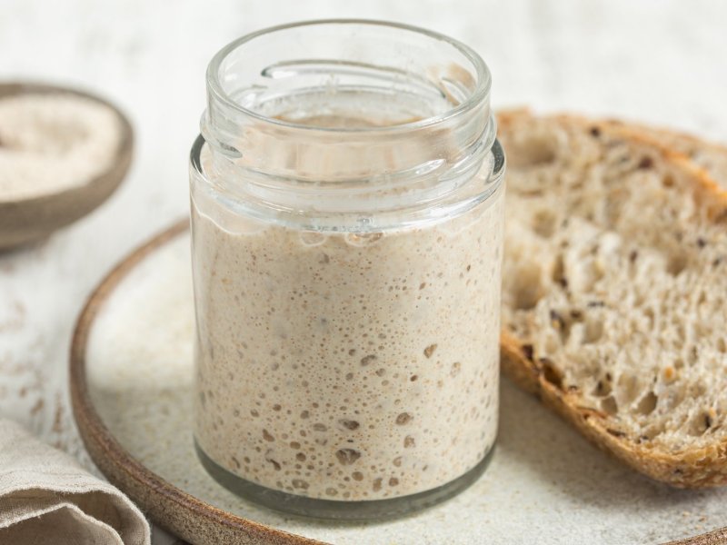 Ist Sauerteig glutenfrei? Ein Glas mit Sauerteigansatz neben einer Scheibe Brot.