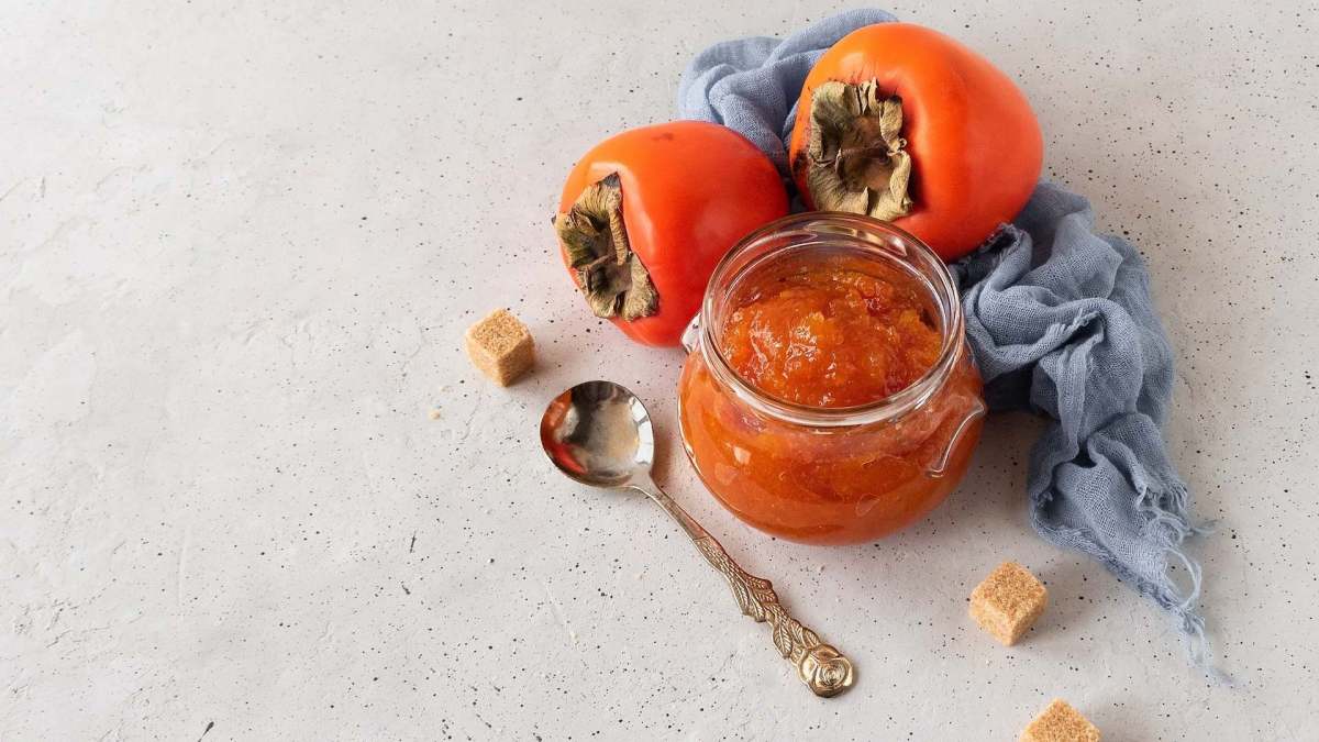 Ein Glas mit Kaki-Marmelade, daneben Zuckerwürfel und ganze Früchte.