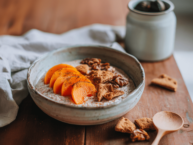 Eine Schale Kaki Spekulatius Porridge auf einem Holztisch. Rechts daneben liegen einige Spekulatiuskeks-Stücke und ein Löffel. Links liegt ein Küchentuch. Im Hintergrund ist ein kleiner Keramikbehälter zu sehen.
