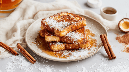 Drei Scheiben Kokos-French-Toast liegen gestapelt auf einem Teller. Sie sind mit Ahornsirup und Kokosraspeln garniert. Neben dem Teller liegen einige Zimtstangen. Im Hintergrund sieht man eine Tasse Kaffee und eine Kokosnuss.