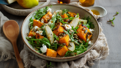 Eine Schale Kürbis-Feta-Salat auf einem beigen Tuch auf einem grauen Holztisch. Links neben der Schüssel liegt ein Holzlöffel. Am oberen Bildrand sieht man eine Birne und mehrere Dressingzutaten.