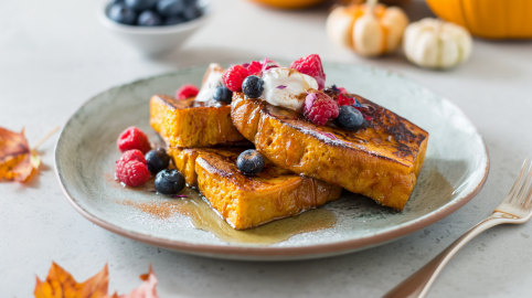 Drei Scheiben Kürbis-French-Toast liegen auf einem rustikalen Keramikteller. Sie sind mit Ahornsirup übergossen und mit Beeren und einem Klecks Joghurt garniert. Im Hintergrund sieht man ein paar Kürbisse, vor und neben dem Teller liegen zwei Blätter.