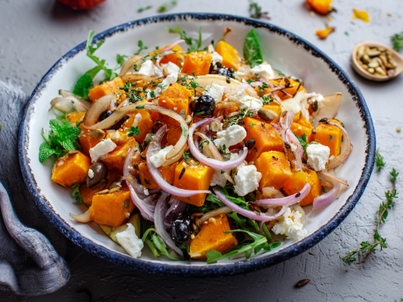 Ein Teller mit Kürbis-Hüttenkäsesalat auf grauem Untergrund. Links neben dem Teller liegt ein Tuch. Drumherum sind einige Frische Kräuter verteilt.