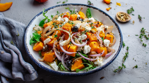 Ein Teller mit Kürbis-Hüttenkäsesalat auf grauem Untergrund. Links neben dem Teller liegt ein Tuch. Drumherum sind einige Frische Kräuter verteilt.