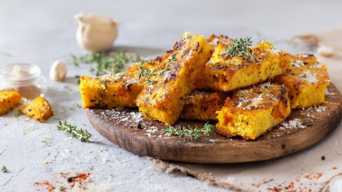 Ein rundes Holzbrett mit einigen Kürbis-Polenta-Schnitten, die aufeinander gestapelt sind. Sie sind mit geriebenem Parmesan und frischem Thymian garniert. Ringsum um und im Hintergrund kann man ein paar Kürbisstücke, eine Knoblauchknolle sowie frische Kräuter und Gewürze verstreut liegen sehen.