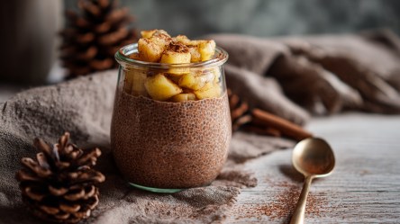 Ein kleines Glas mit Lebkuchen-Chiapudding, der mit Apfelkompott getoppt ist. Ringsum kann man Tannenzapfen, Zimtstangen und ein drapiertes Tuch erkennen.