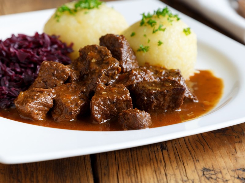 Malzbiergulasch mit Knödeln und Rotkohl auf einem weißen Teller.