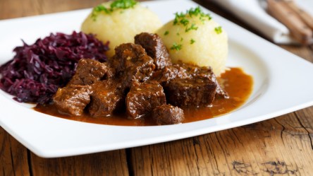 Malzbiergulasch mit Knödeln und Rotkohl auf einem weißen Teller.