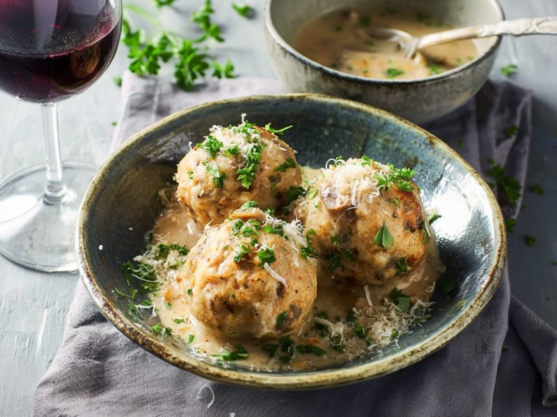 Ein Teller mit drei Maronen-Knödel, die mit Petersilie und Parmesan garniert ist. Dabei steht ein Glas Rotwein und eine Schale mit Rahmsoße.