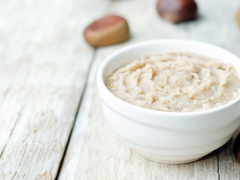 Eine Schale Maronenbutter auf einem hellen Holzuntergrund. Drum herum sind Maronen.