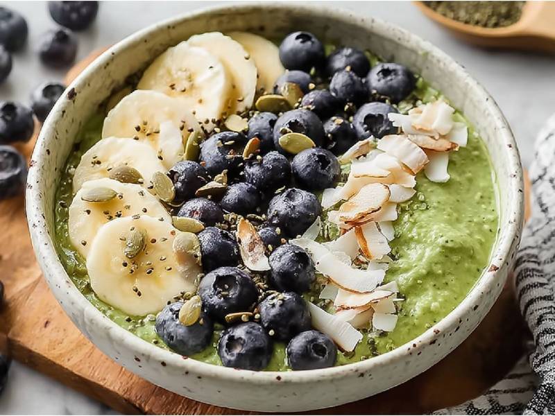 Schale mit Matcha-Overnight-Oats mit Kokos, Bananenscheiben, Kokoschips, Heidelbeeren und Kürbissamen als Topping