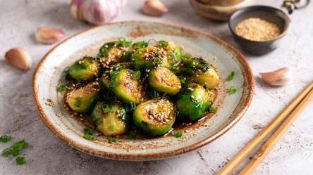 Ein Teller mit Miso-Rosenkohl. Ringsum liegen Stäbchen, Knoblauchzehen und eine Schale mit Soße.