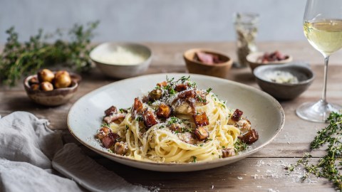 Ein Teller Pasta mit Maronen und Speck. Das Ganze ist mit geriebenem Parmesan und frischen Thymianblättchen garniert. Im Hintergrund steht ein Glas Weißwein und es stehen Schalen mit Zutaten herum.
