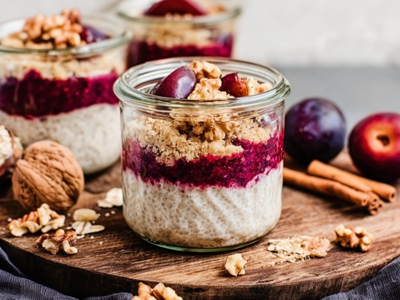 Mehrere kleine Gläser mit Pflaumen-Crumble-Chiapudding, die auf einem Holzbrett platziert sind. Außerdem liegen Zimtstangen, einige Walnüsse und Pflaumen herum.