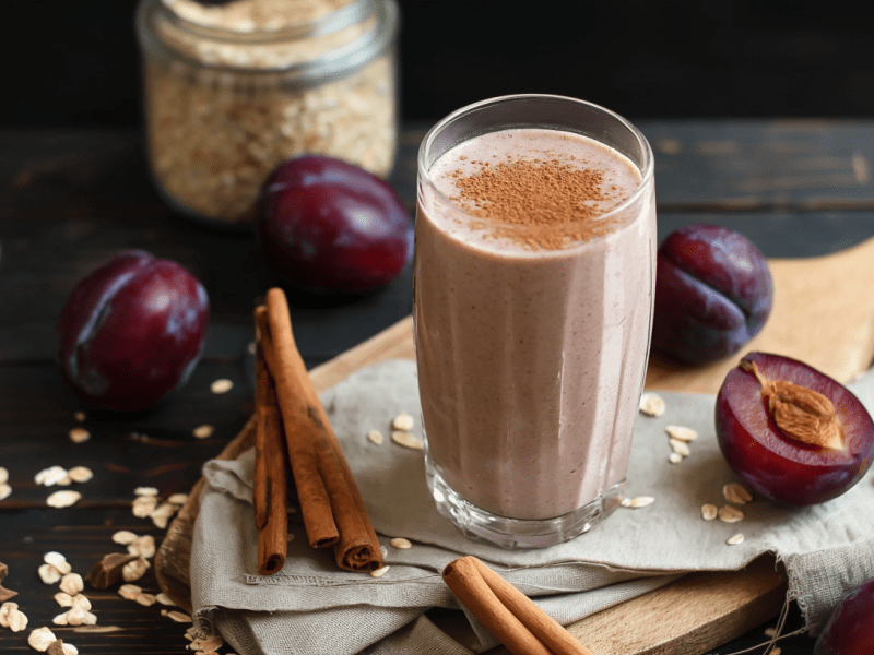 Ein Pflaumen-Dattel-Shake auf einem Holzbrett. Um den Shake liegen mehrere Pflaumen und Zimtstangen verteilt. Im Hintergrund kann man ein Glas mit Haferflocken erkennen.