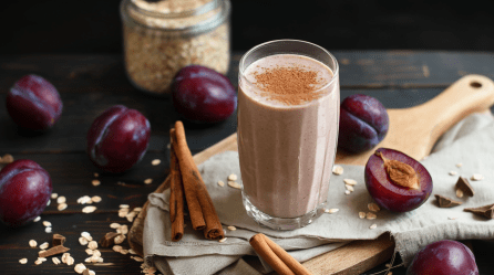 Ein Pflaumen-Dattel-Shake auf einem Holzbrett. Um den Shake liegen mehrere Pflaumen und Zimtstangen verteilt. Im Hintergrund kann man ein Glas mit Haferflocken erkennen.