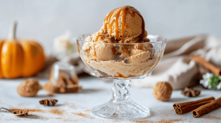 Eine Schale mit Pumpkin-Spice-Eis. Um die Schale verteilt ist Zimt verteilt. Im Hintergrund sieht man einen Kürbis sowie diverse Gewürze.