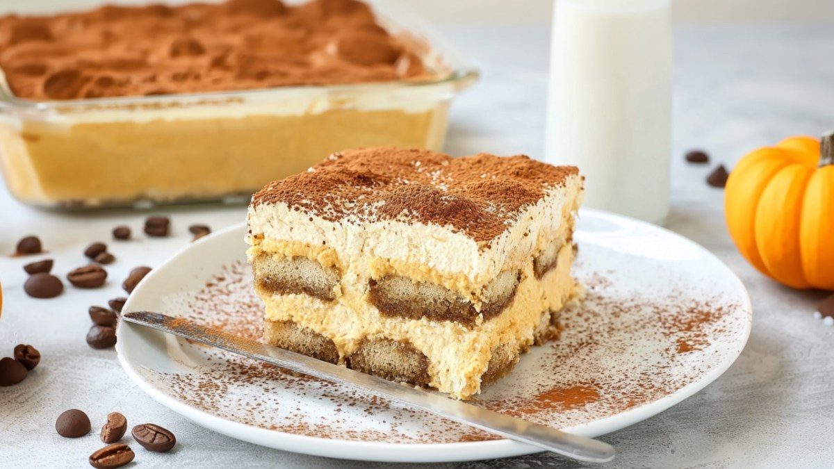 Ein Stück Pumpkin Spice Tiramisu auf einem Teller. Im Hintergrund ist das ganze Tiramisu zu sehen.