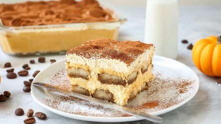 Ein Stück Pumpkin Spice Tiramisu auf einem Teller. Im Hintergrund ist das ganze Tiramisu zu sehen.