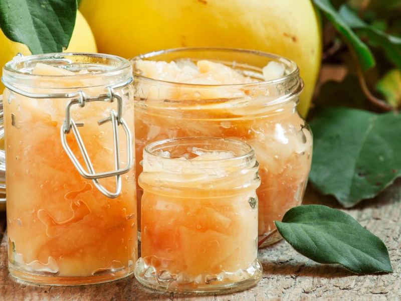 Quitten einkochen: Mehrere Gläser mit eingemachten Früchten, dahinter rohe Quitten.