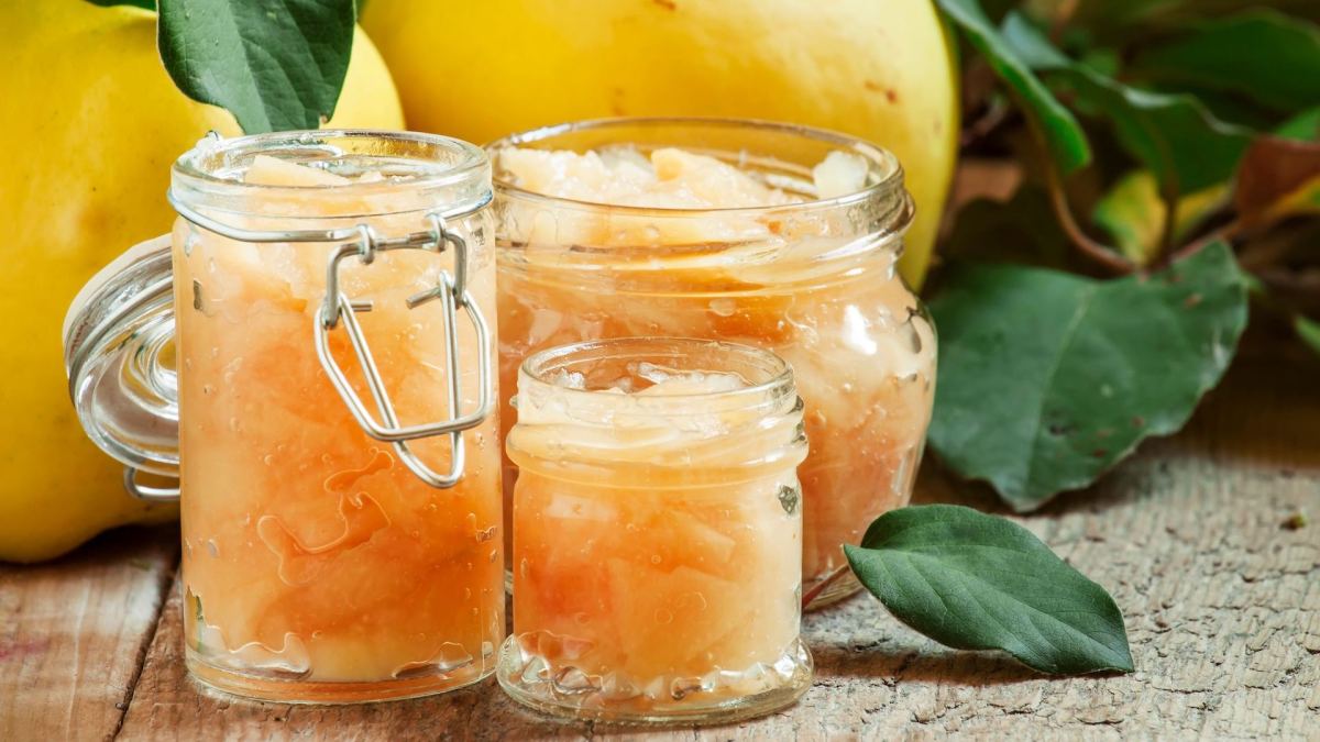 Quitten einkochen: Mehrere Gläser mit eingemachten Früchten, dahinter rohe Quitten.