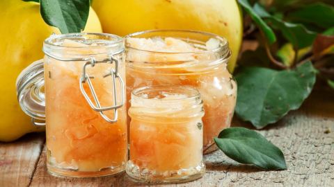 Quitten einkochen: Mehrere Gläser mit eingemachten Früchten, dahinter rohe Quitten.