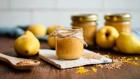 Ein offenes Glas mit Quitten-Senf. Daneben liegt ein Holzlöffel mit gelben Senfkörnern. Ringsum liegen Quitten sowie Orangenabrieb und Senfkörner verteilt. Im Hintergrund sind weitere Gläser mit Quitten-Senf erkennbar.