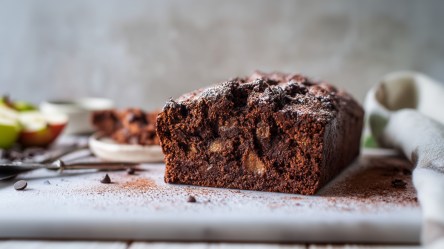 Ein Schokoladenkuchen mit Äpfeln, der auf einer hellen Arbeitsfläche liegt. Der Kuchen ist angeschnitten. Im Hintergrund kann man aufgeschnittene Äpfel erahnen, genauso wie eine Schale mit Kakao, Vanilleschoten und Schokotropfen.