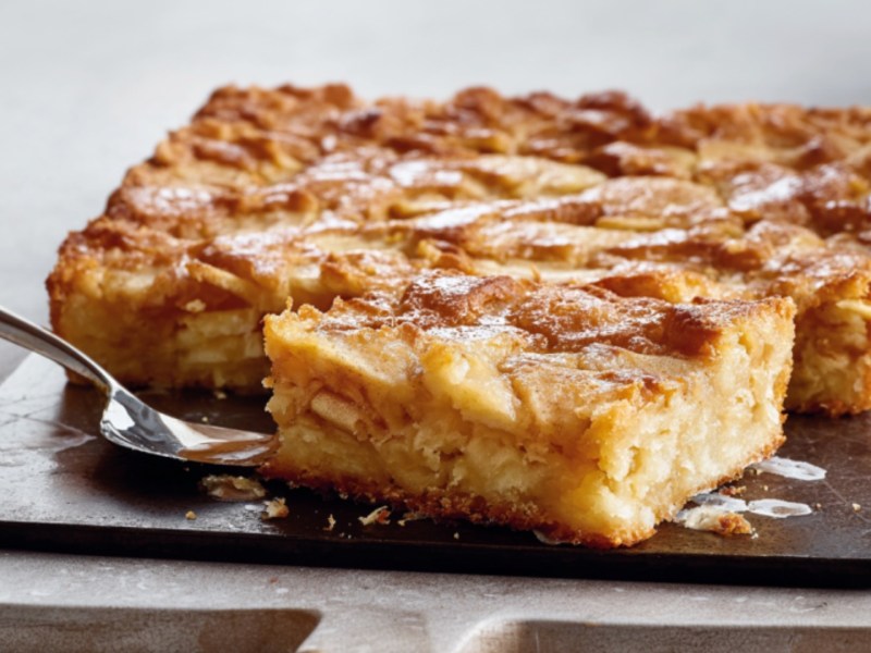 Ein Stück schwedischer Apfelkuchen vom Blech liegt vor dem restlichen Kuchen auf einer Platte.