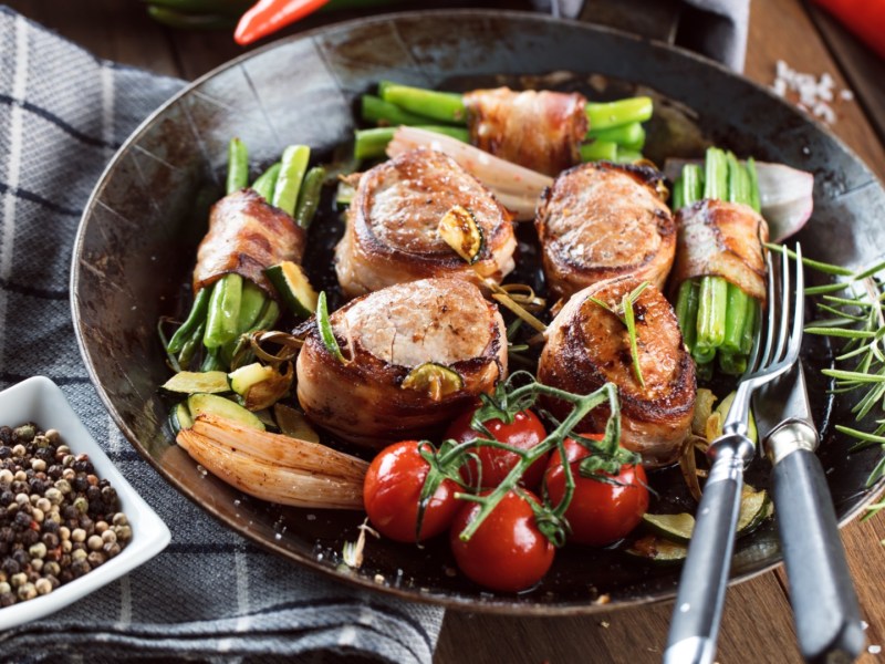 Schweinefilet im Speckmantel mit Ofengemüse und Bohnen im Speckmantel auf einem Teller.