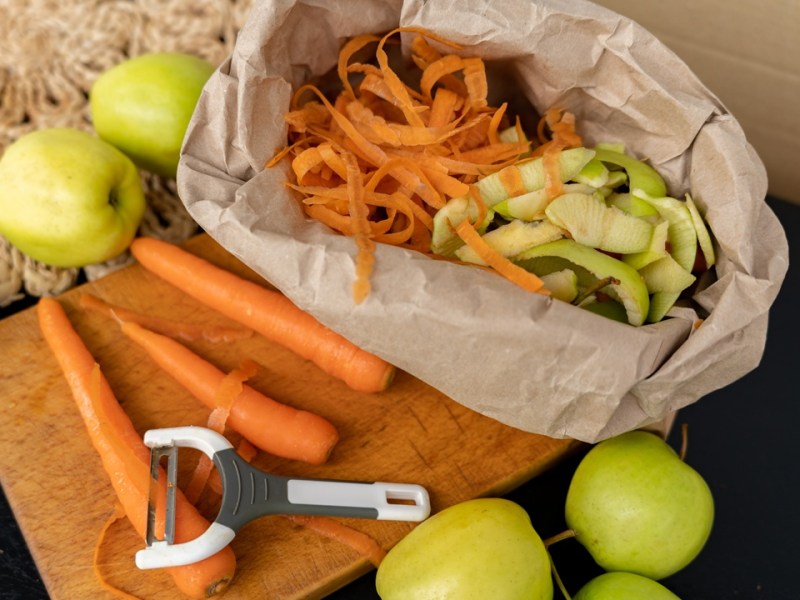 Eine Papiertüte mit Gemüseabfällen, daneben liegen geschälte Karotten und Äpfel. Ein Sparschäler ist ebenfalls zu sehen.