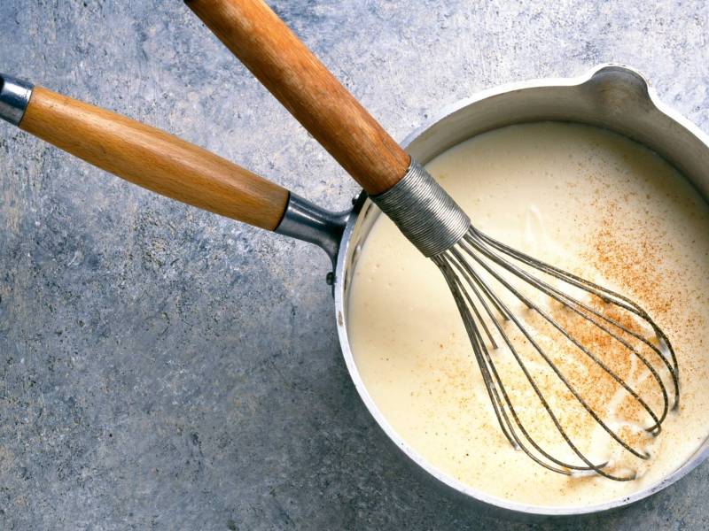 Draufsicht: Ein Topf mit heller Soße, in dem ein Schneebesen liegt.