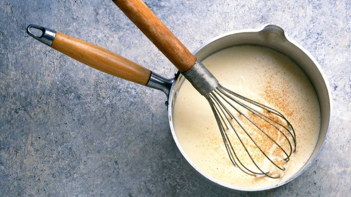 Draufsicht: Ein Topf mit heller Soße, in dem ein Schneebesen liegt.