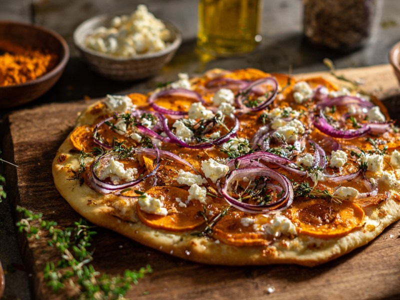 Ein Süßkartoffel-Flammkuchen, der auf einem Holzbrett liegt. Ringsum sind frische Zutaten zu sehen.