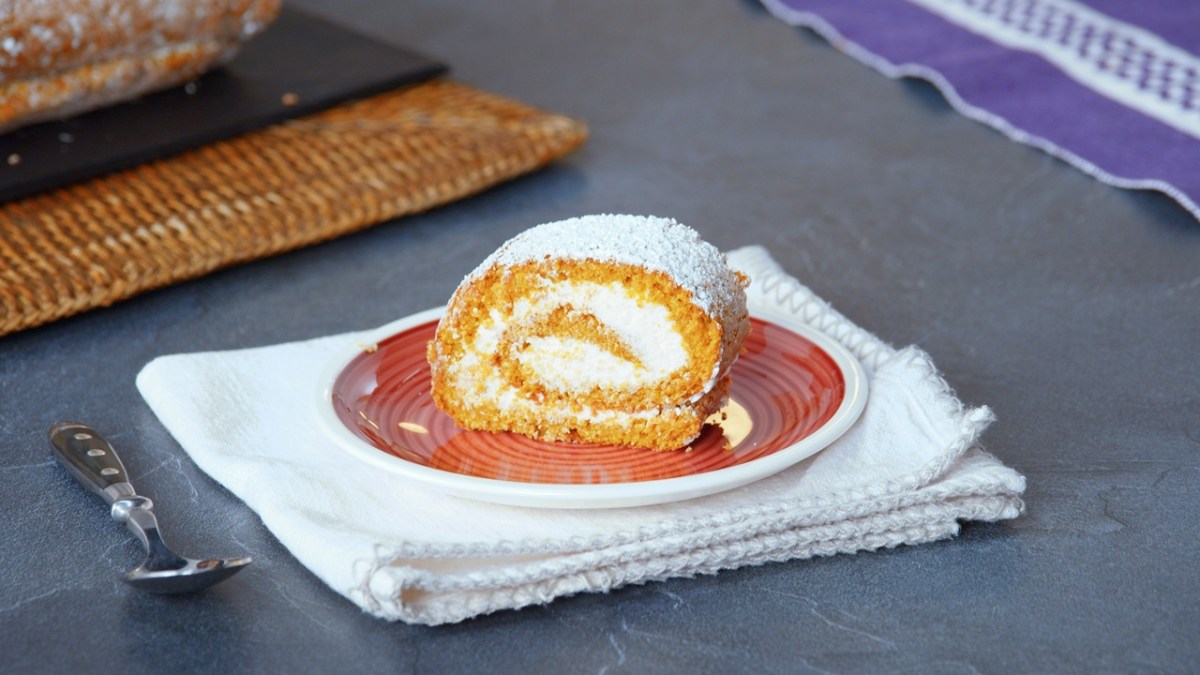 Ein Stück Kürbisrolle, serviert auf einem Teller. Der Teller steht auf einer weißen Stoffserviette, daneben liegt eine Kuchengabel.